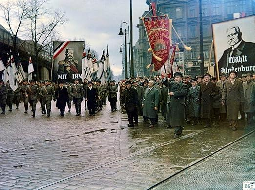 Eine Parade mit Bannern und Flaggen, Soldaten marschieren auf einer nassen Straße. Porträts von Führern sind sichtbar. Menschen schauen zu.