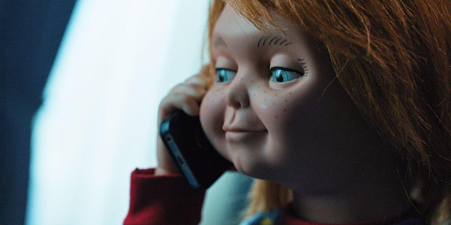 Eine Puppe mit roten Haaren und blauen Augen hält ein Telefon. Der Hintergrund ist verschwommen.