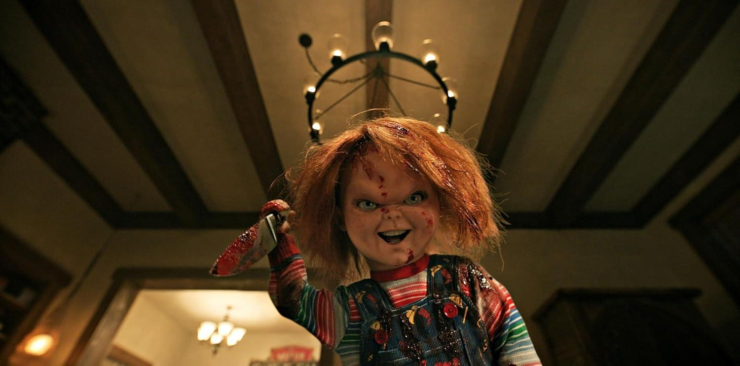 Chucky-Puppe hält ein blutiges Messer. Rotes Haar, blaue Latzhose und ein finsteres Grinsen. Kronleuchter und Holzdecke im Hintergrund.