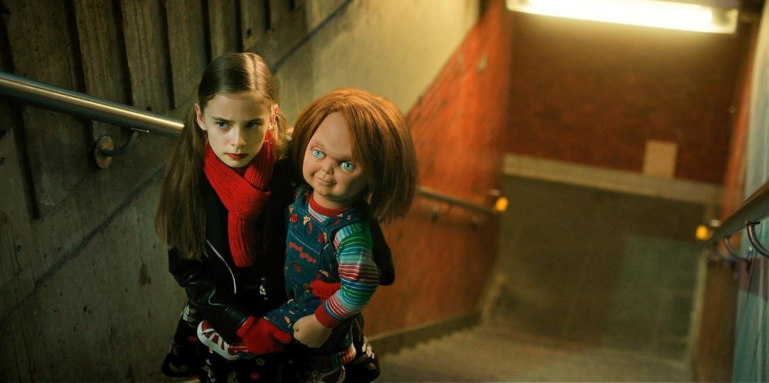 Ein Mädchen hält eine Chucky-Puppe in einem schwach beleuchteten Treppenhaus. Die Puppe hat rotes Haar und blaue Augen. Das Mädchen trägt einen roten Schal.