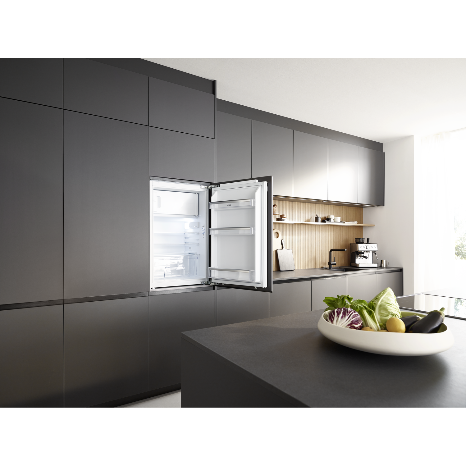 Modern kitchen with open fridge. Vegetables in a bowl on a countertop. Dark gray cabinets and appliances. 