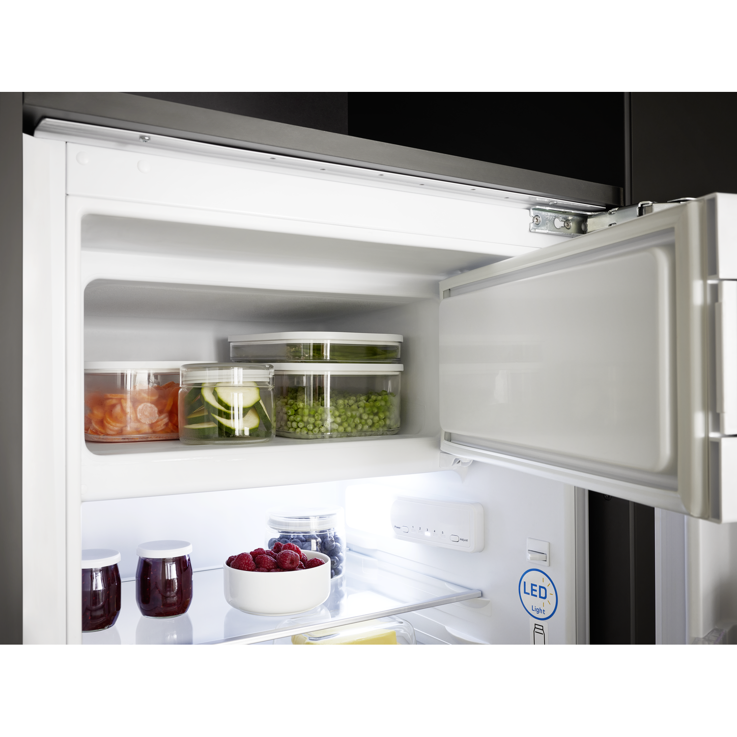 Opened fridge with food: vegetables, berries, and jam. White interior with LED light. Door is open.