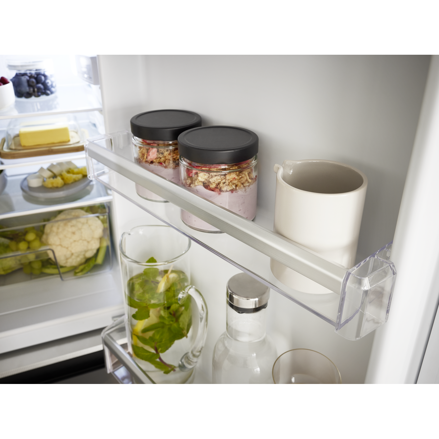 Refrigerator interior with food. Jars with granola, a jug of water with mint and lemon, and other food items.