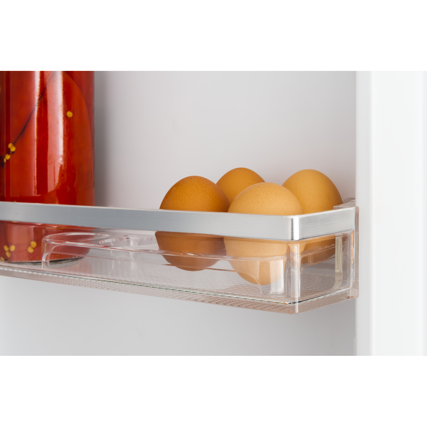 Inside a fridge: brown eggs in a clear container, jar with red contents, all on a shelf. White fridge interior.