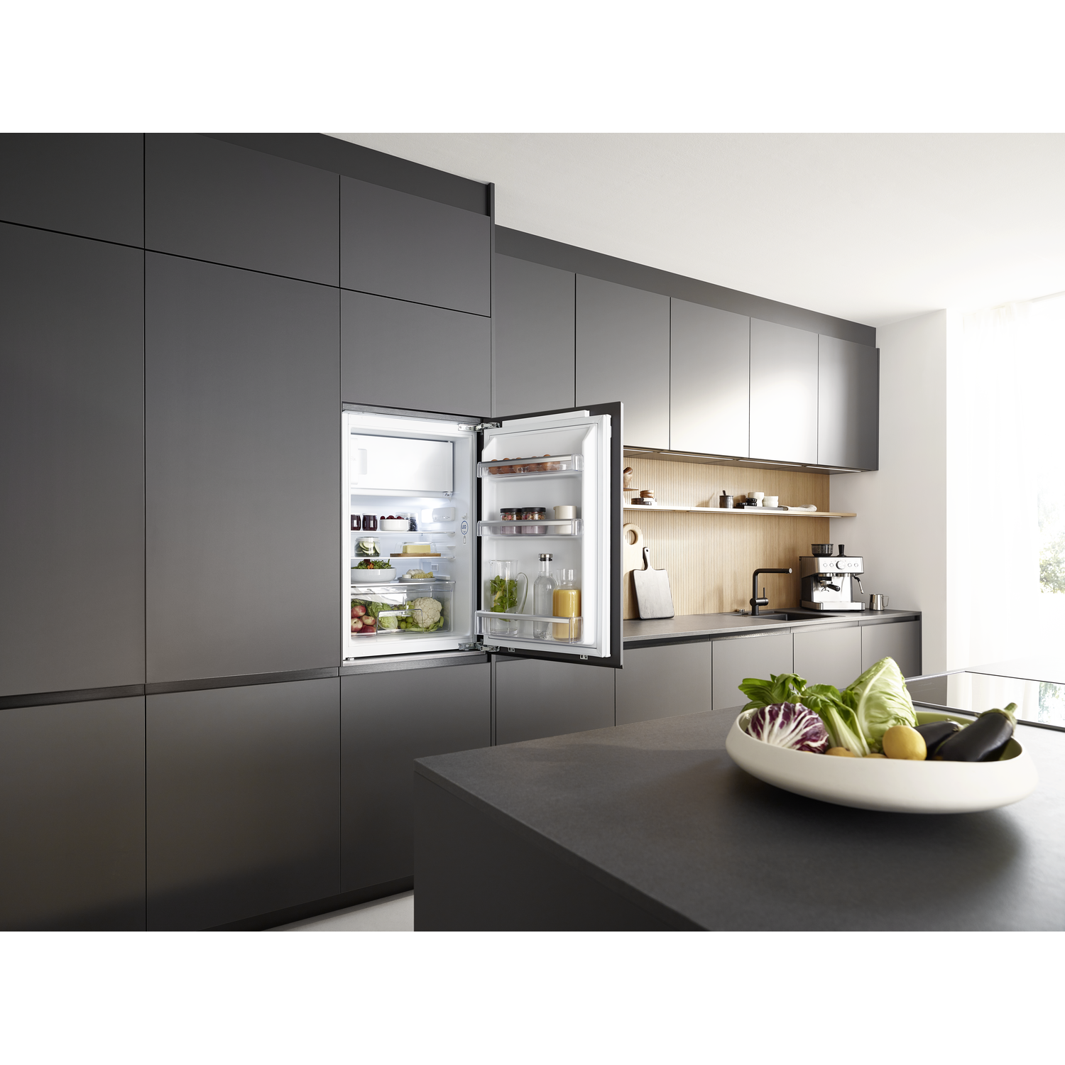 Kitchen scene with open fridge. Vegetables in a bowl on the counter. Dark grey cabinets and countertop. Bright interior.