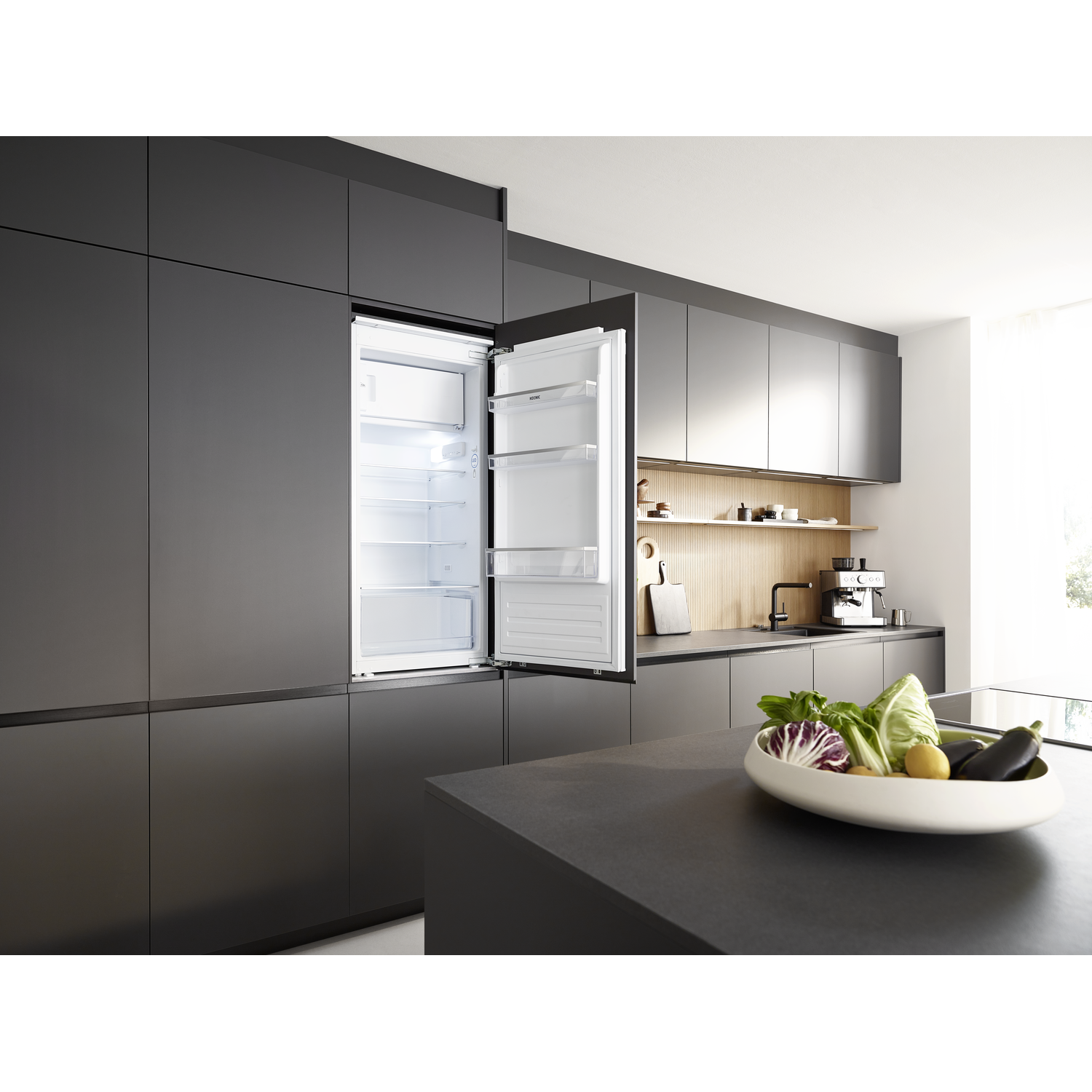 Open fridge in modern kitchen. Vegetables in a bowl on a dark countertop. Dark cabinets and wooden accents.