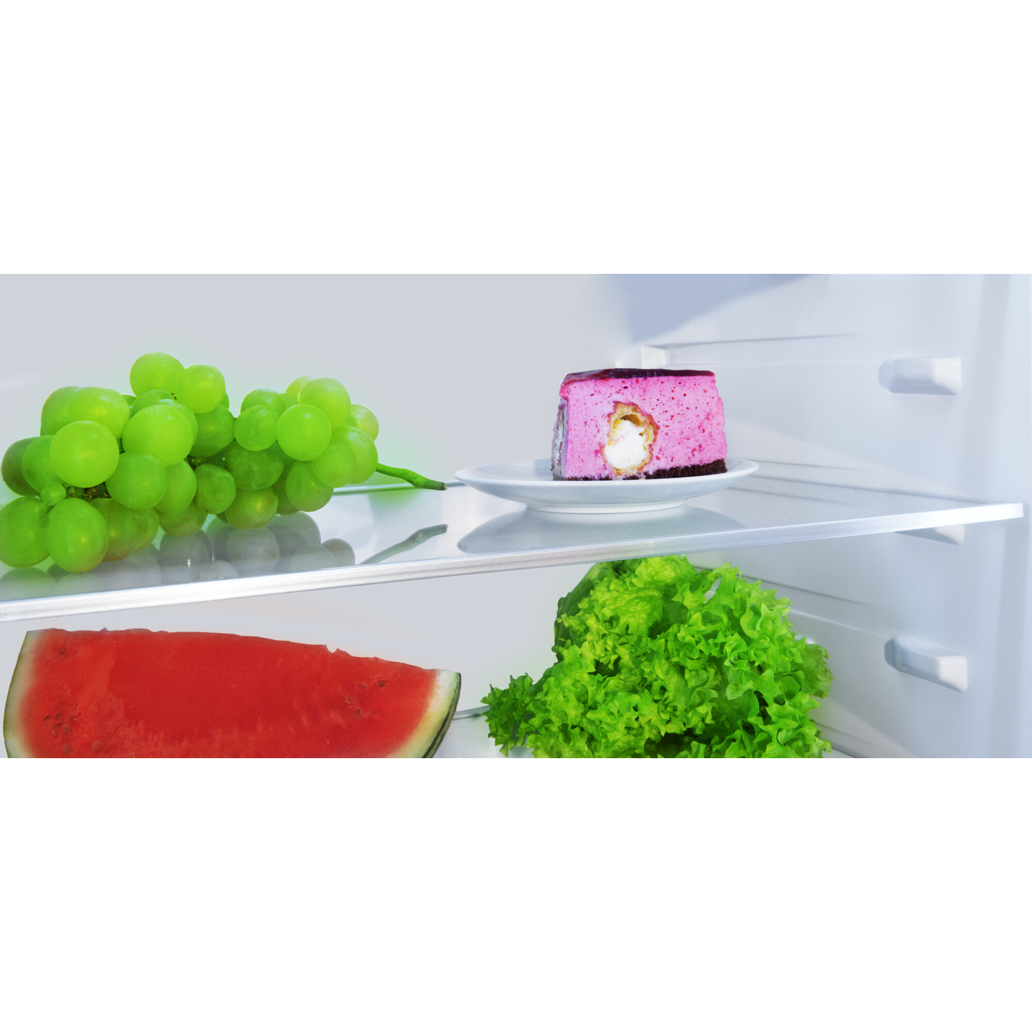 Inside a fridge: grapes, cake on a plate, watermelon, and lettuce. Pink cake, green grapes, and red watermelon are visible.