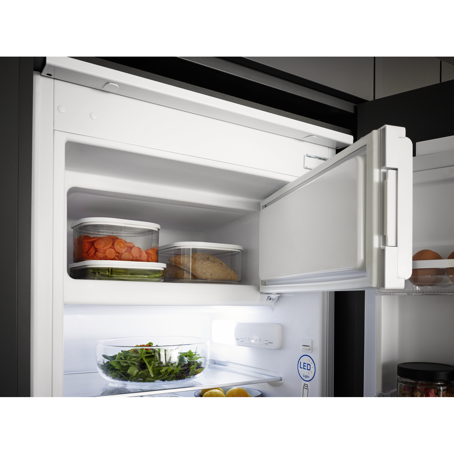 Open refrigerator with food inside: carrots, bread, salad, eggs, and jar. The fridge is white with LED light.
