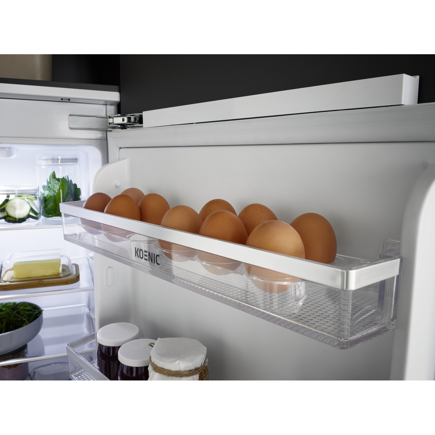 Inside a refrigerator, brown eggs sit in a clear container. Jars, butter, and vegetables are also visible.
