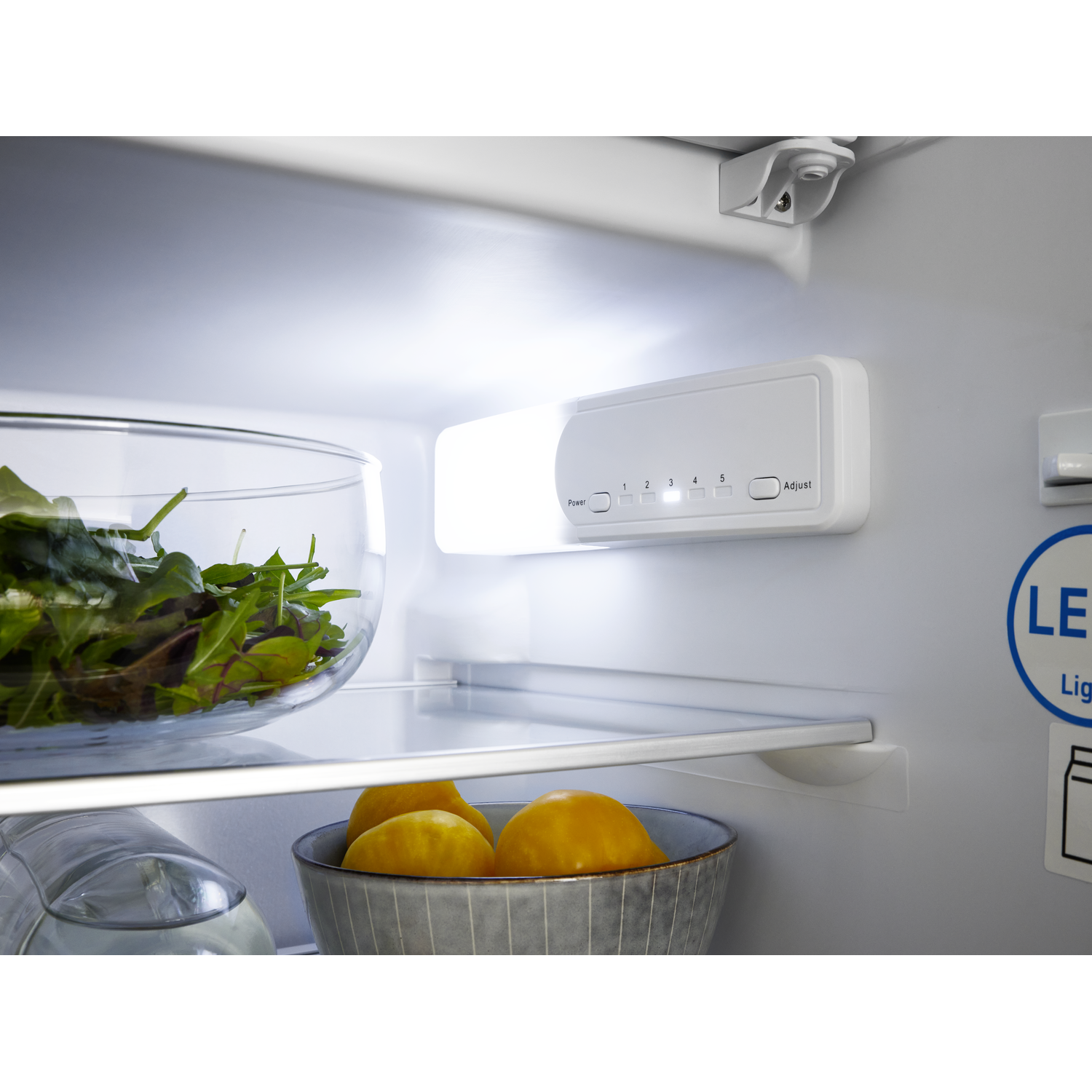 Inside a refrigerator: a bowl of salad, bowl of oranges, shelf, and control panel with lights.