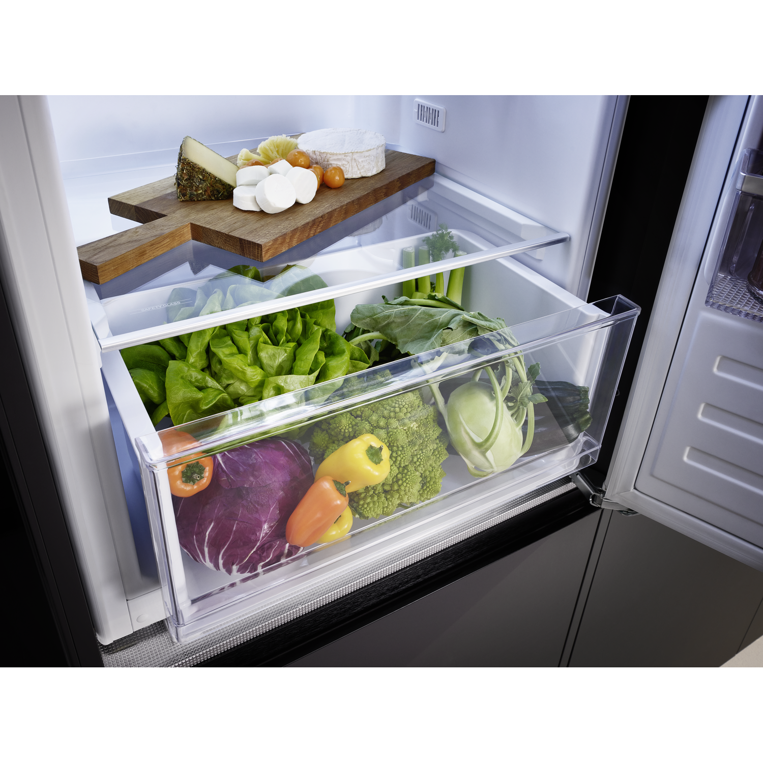 Open fridge with cheeses on a wooden board. Vegetables in a drawer below. Brightly lit interior.