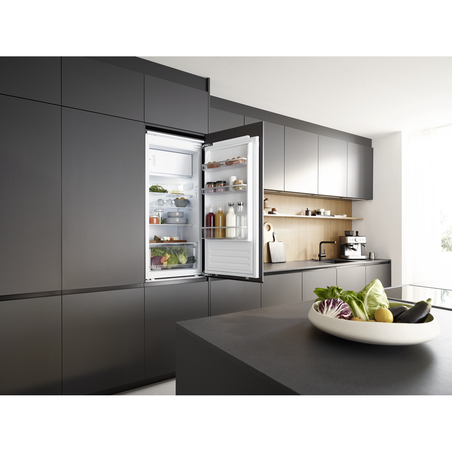 Open refrigerator in a modern kitchen. It contains food. A bowl of vegetables is on the counter. Dark gray cabinets.