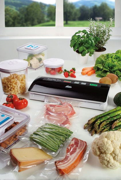 Produkty spożywcze: mięso, warzywa, truskawki, makaron w pojemnikach. Zgrzewarka próżniowa na białej powierzchni. Zielone wzgórza i okno w tle.