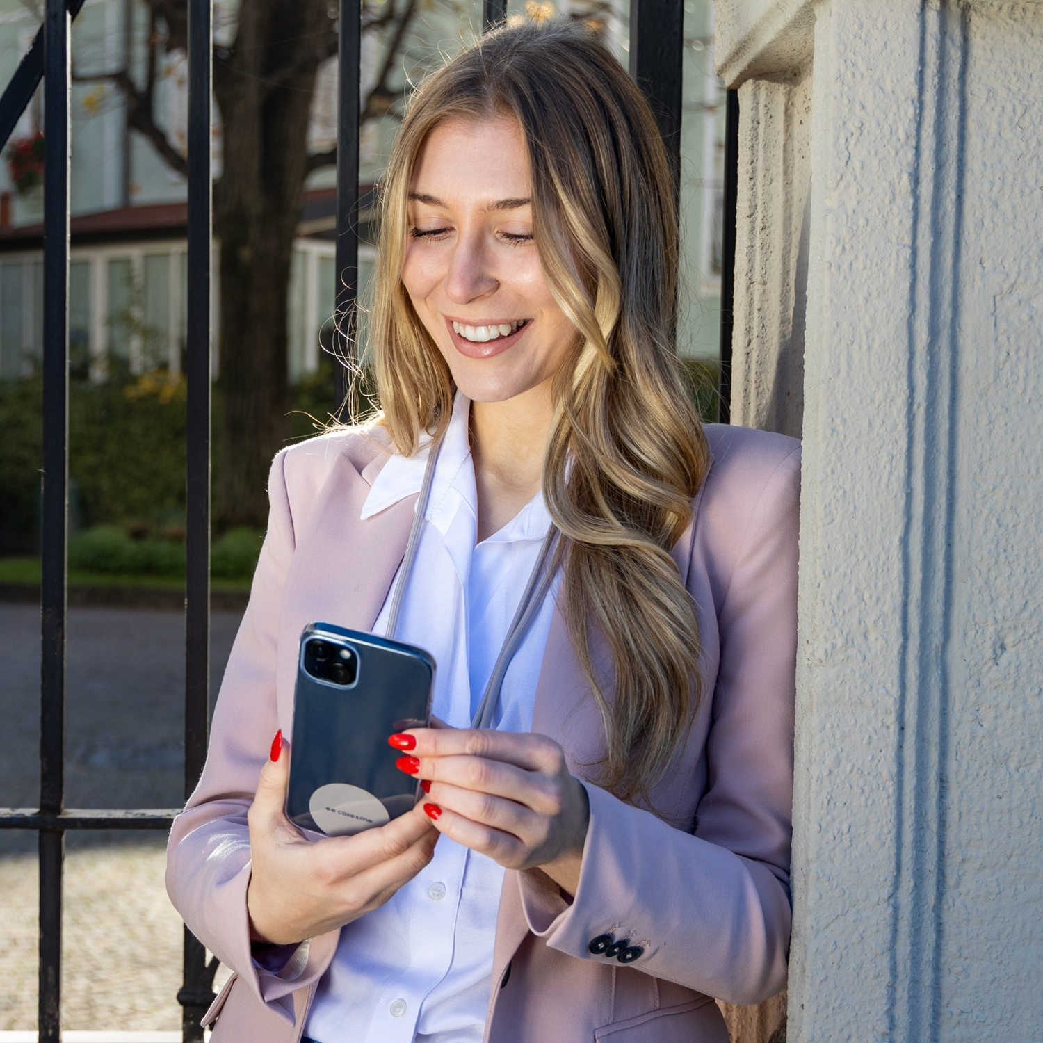 Donna in blazer rosa sorride, tiene un telefono. Unghie rosse. Appoggiata a un muro bianco vicino a una recinzione nera. Giorno soleggiato.