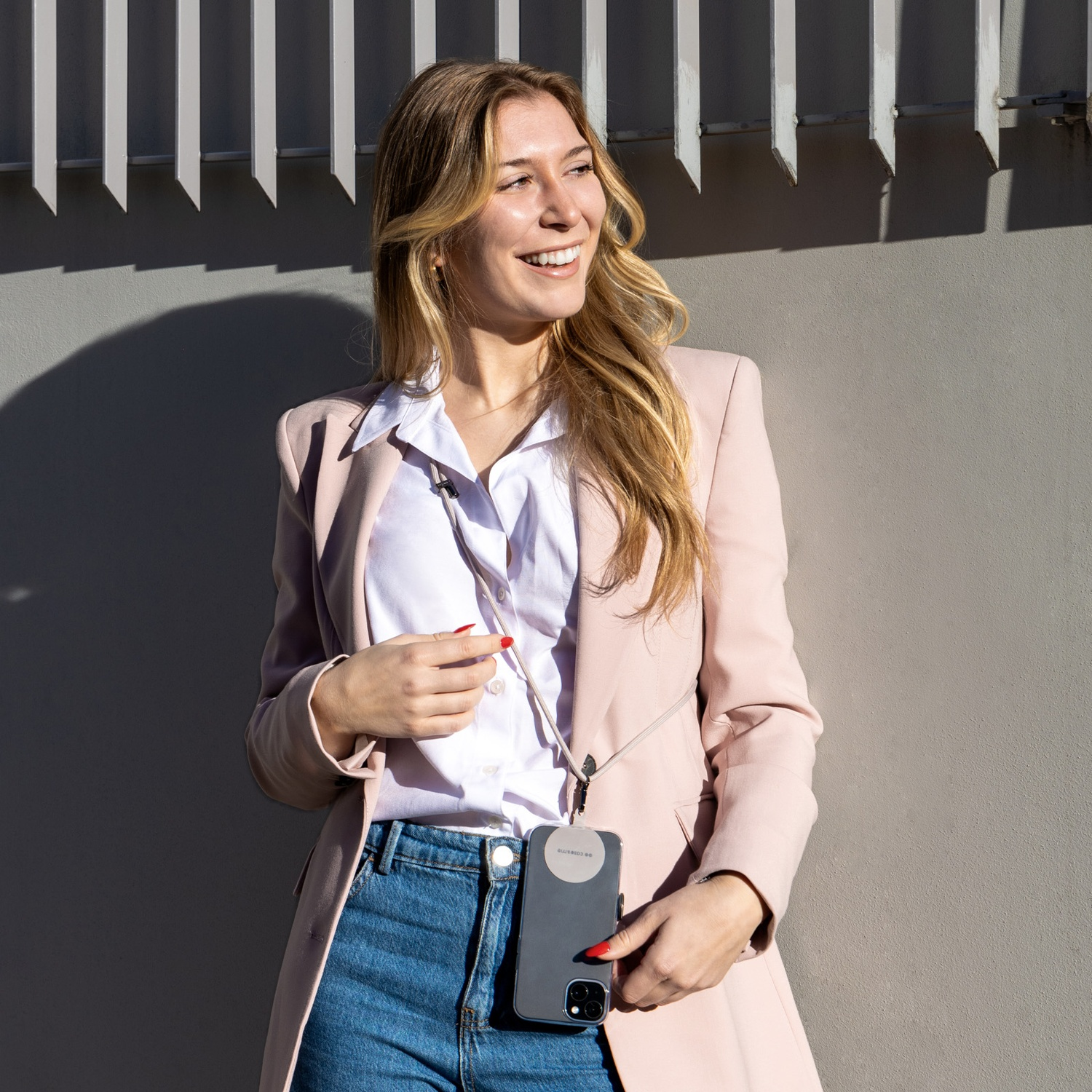 Donna in blazer sorride, tiene in mano un telefono con cordino. Muro grigio e barre metalliche sullo sfondo.
