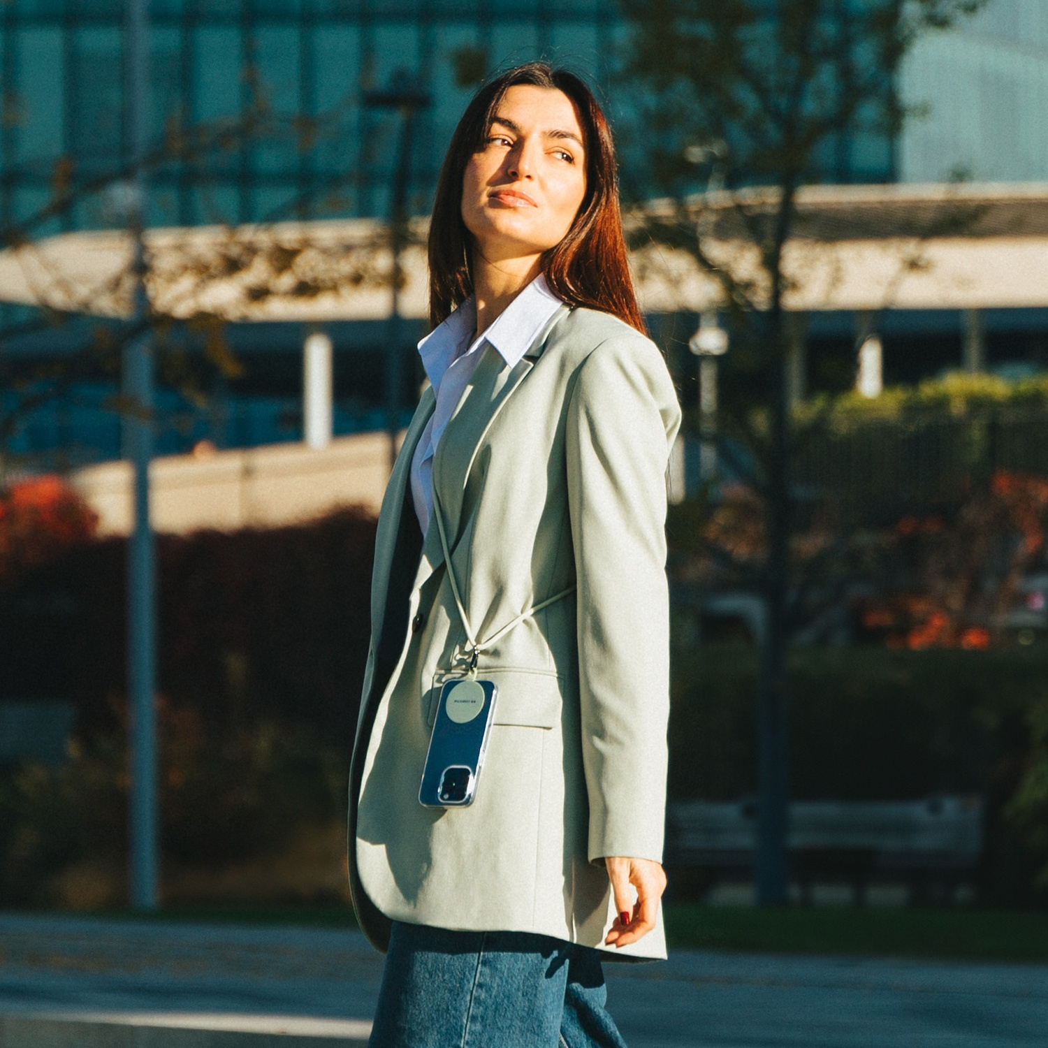 Donna in blazer cammina all'aperto, telefono con cordino. Sfondo con alberi ed edificio.