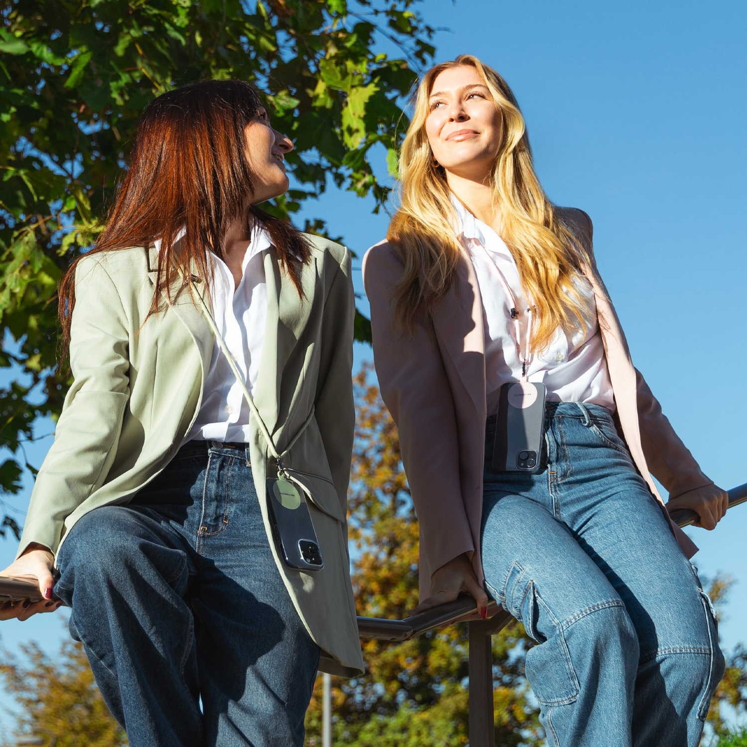 Due donne sedute su una ringhiera, che guardano in alto. Una ha i capelli rossi, l'altra biondi. Indossano blazer e jeans, con custodie per telefoni.