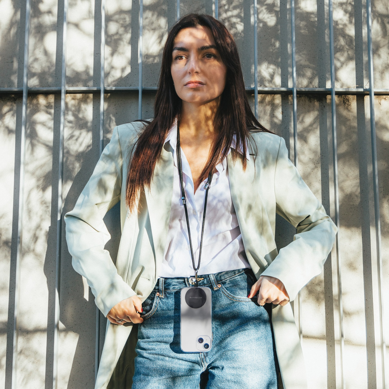 Una donna in blazer e jeans è in piedi vicino a una griglia metallica. Ha una collana con custodia per telefono. La luce solare proietta ombre.