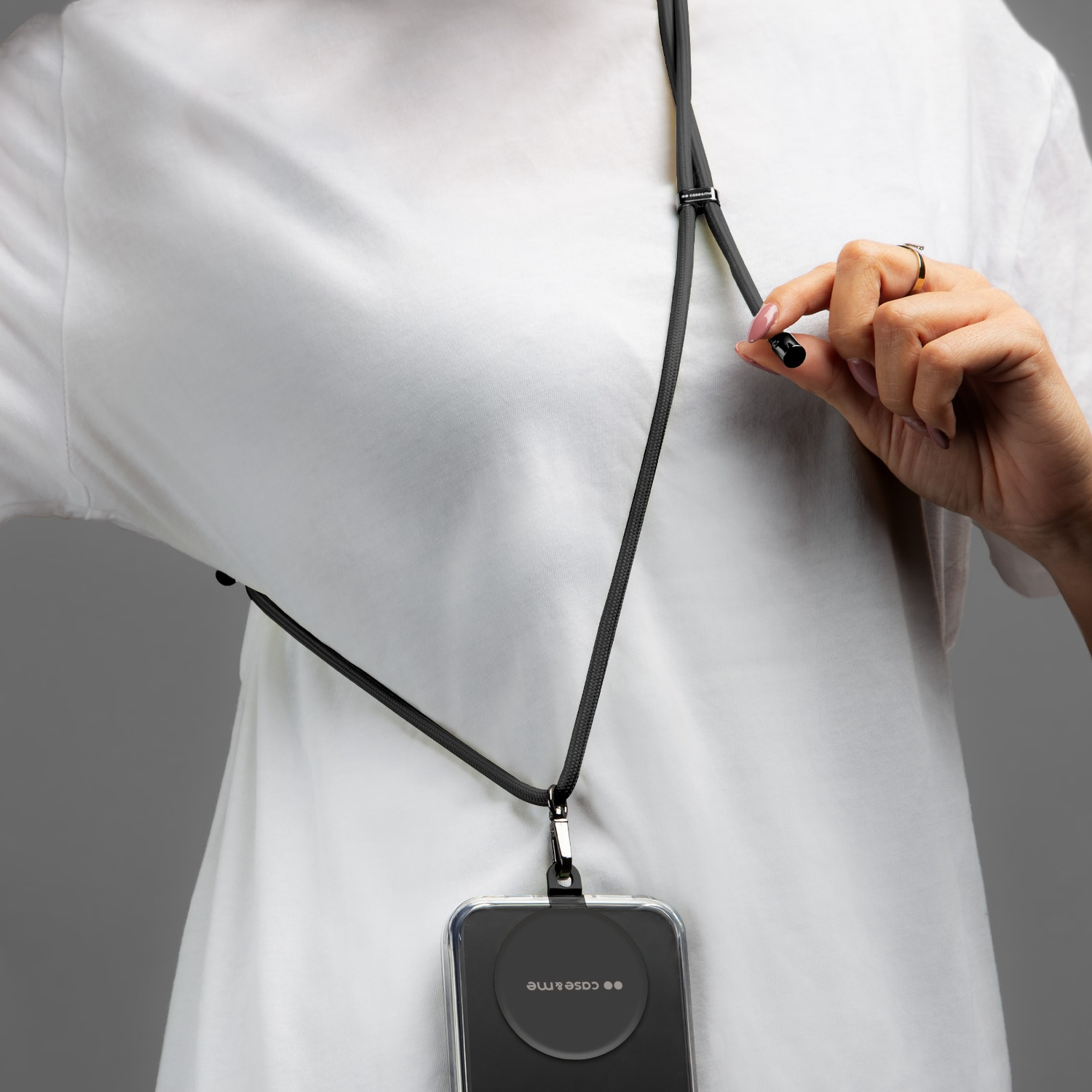 Persona con maglietta bianca tiene un cordino nero con custodia del telefono. Anello al dito. Sfondo grigio.