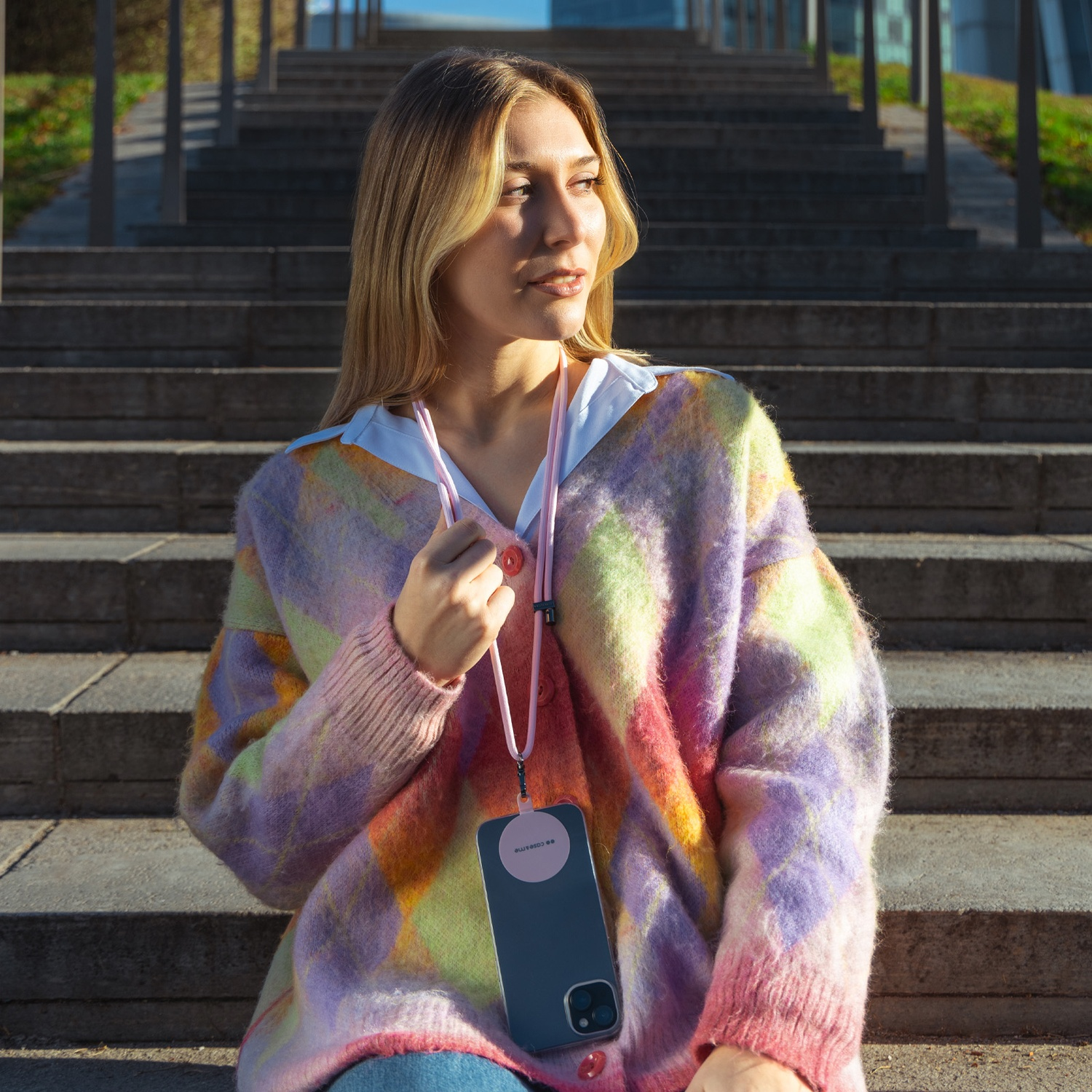 Donna bionda con maglione colorato, tiene un telefono con cinturino rosa, seduta su gradini di cemento.