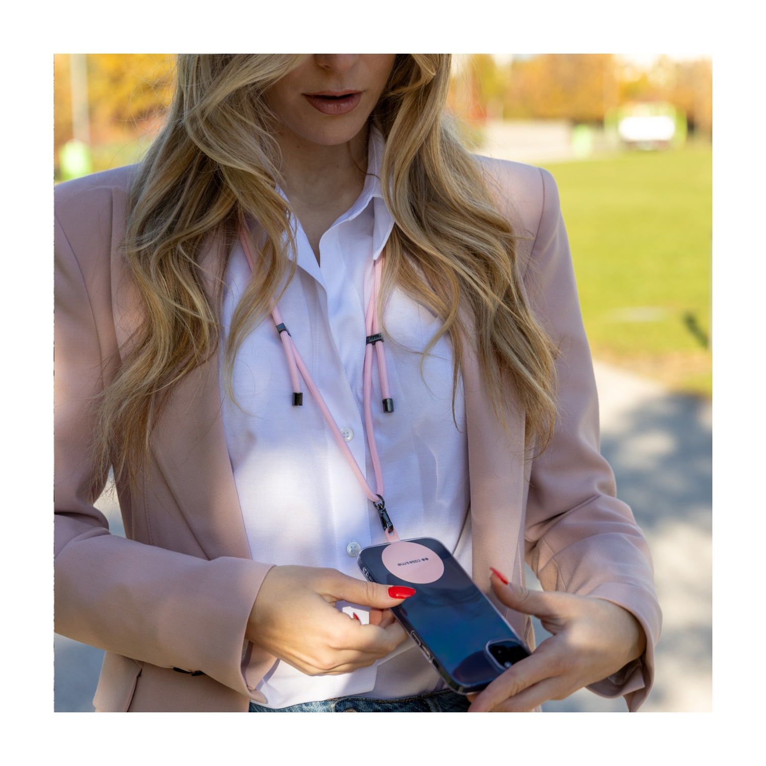 Donna con un cordino rosa che tiene un telefono. Indossa un blazer, camicia bianca. Lo sfondo è un parco con colori autunnali.