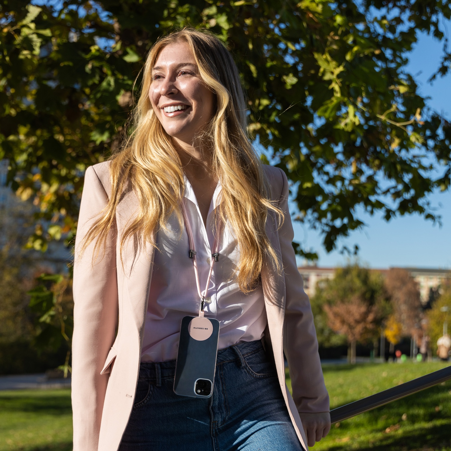 Donna bionda sorride, indossa un blazer rosa e jeans, tiene in mano un telefono con un cordino. Alberi e cielo blu sullo sfondo.