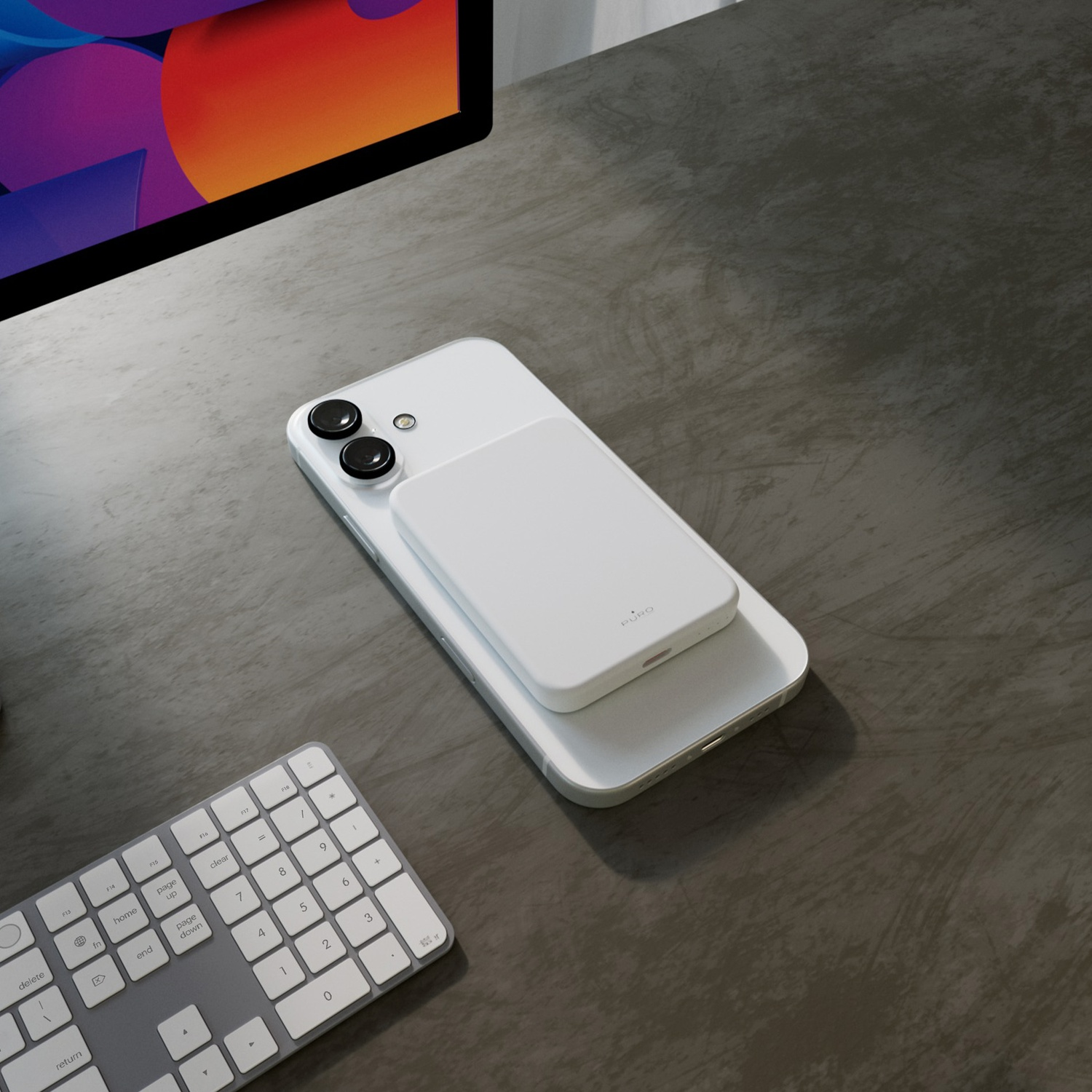 Un telefono bianco con un caricabatterie bianco su una scrivania grigia. Sono visibili anche una tastiera e uno schermo.