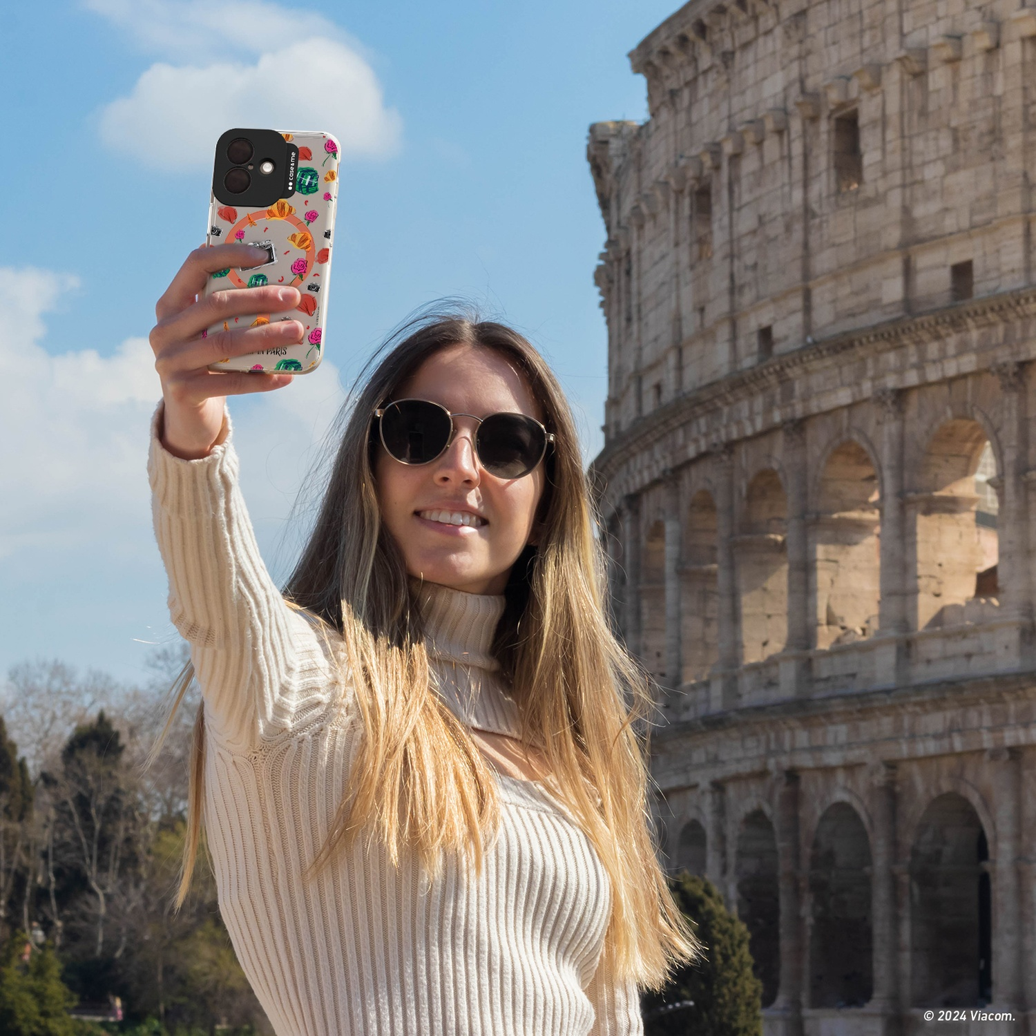 Donna che fa un selfie davanti al Colosseo. Indossa occhiali da sole e un maglione color crema.