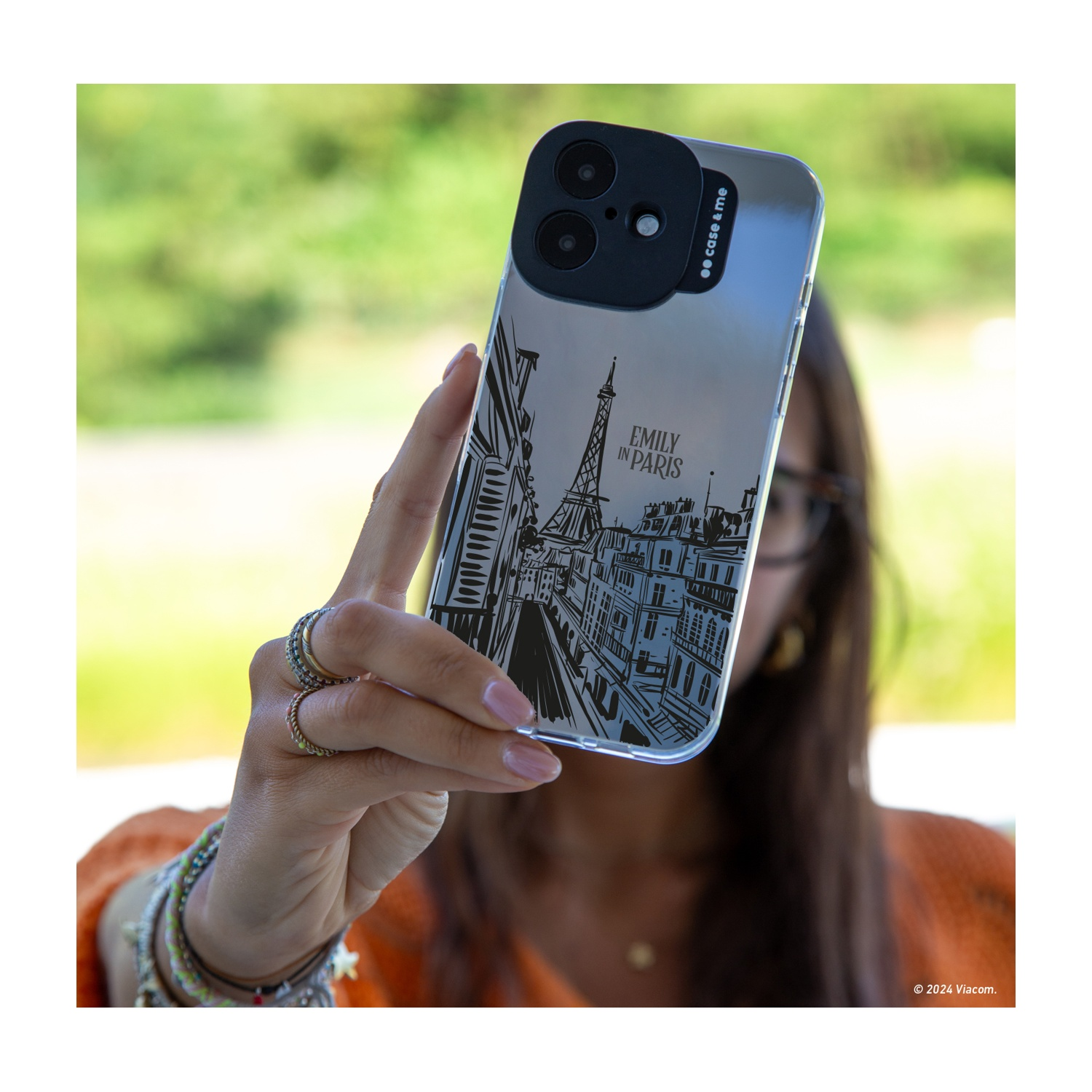 Una donna tiene in mano un telefono con custodia "Emily in Paris", design Torre Eiffel. Anelli e braccialetti visibili. Sfondo verde sfocato.