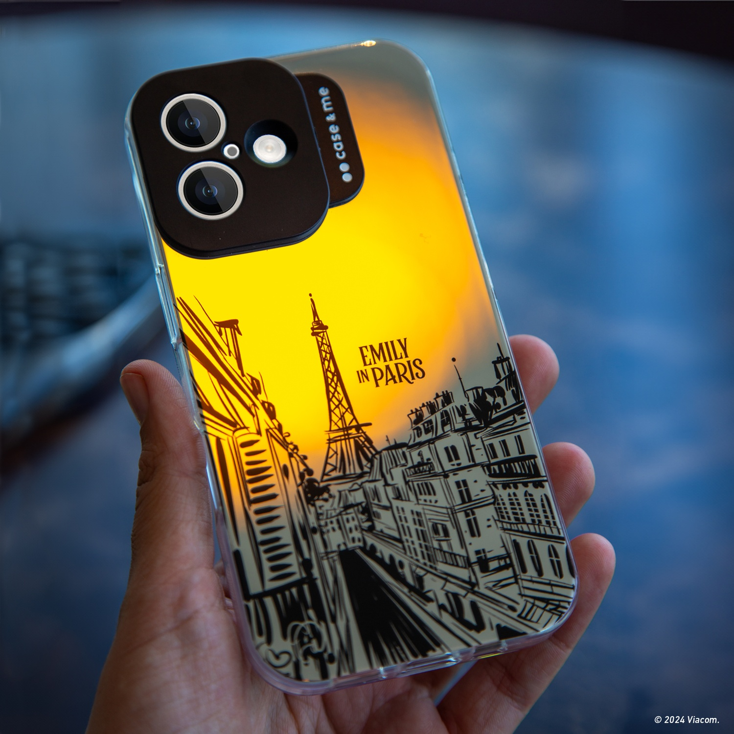 Mano che tiene un telefono con custodia "Emily in Paris". La custodia mostra la Torre Eiffel e gli edifici urbani su sfondo giallo.