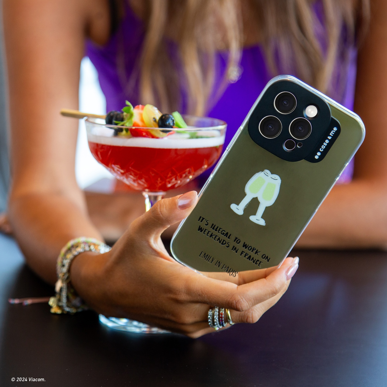 Donna tiene telefono con cocktail. La custodia dice "Emily in Paris". Un cocktail con frutta è davanti.