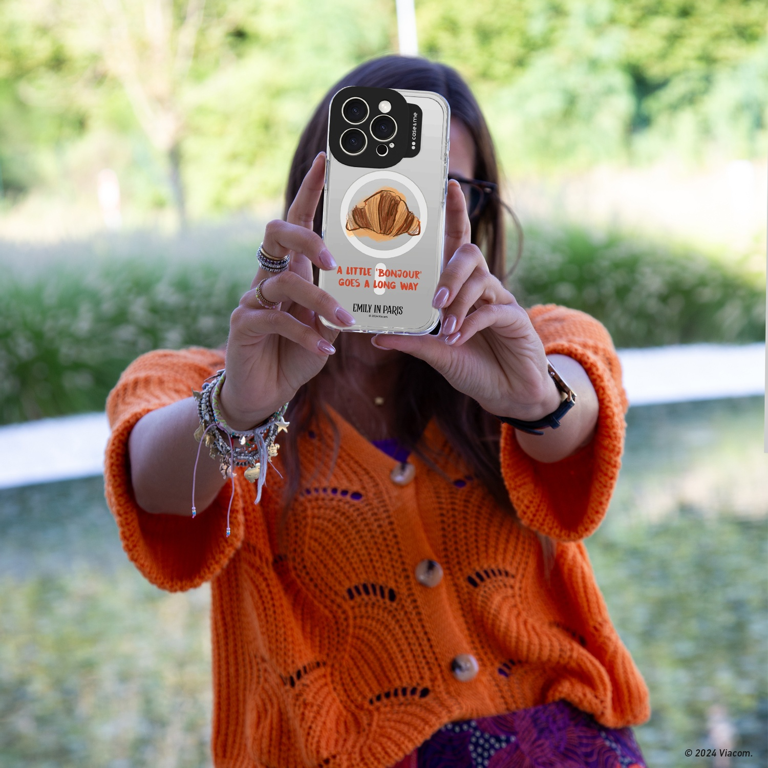 Donna che tiene la custodia del telefono con croissant e testo. Indossa un maglione arancione. Sfondo verde.