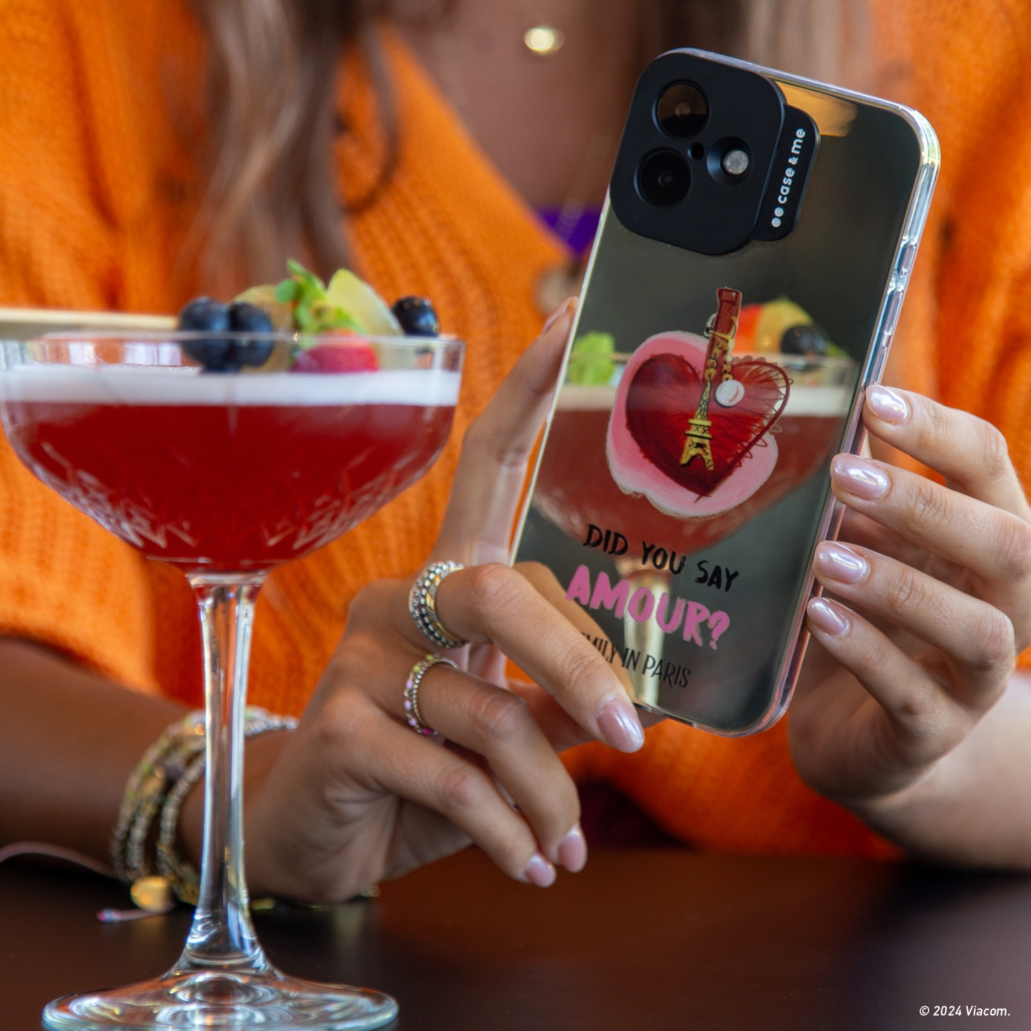 Donna che tiene in mano un telefono con un design di Parigi. Un cocktail con frutta è di fronte. Indossa anelli e braccialetti.