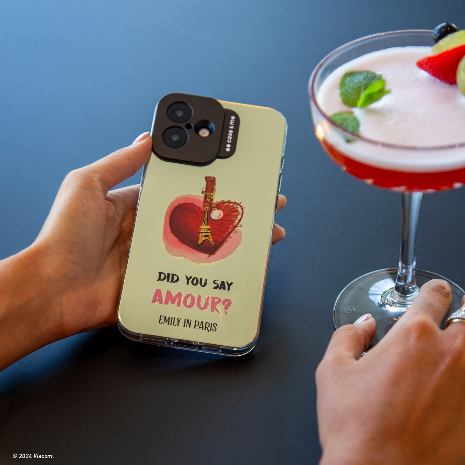 Mano che tiene custodia del telefono con design Torre Eiffel, bicchiere da cocktail vicino. Custodia dice 'Did you say Amour? Emily in Paris.'