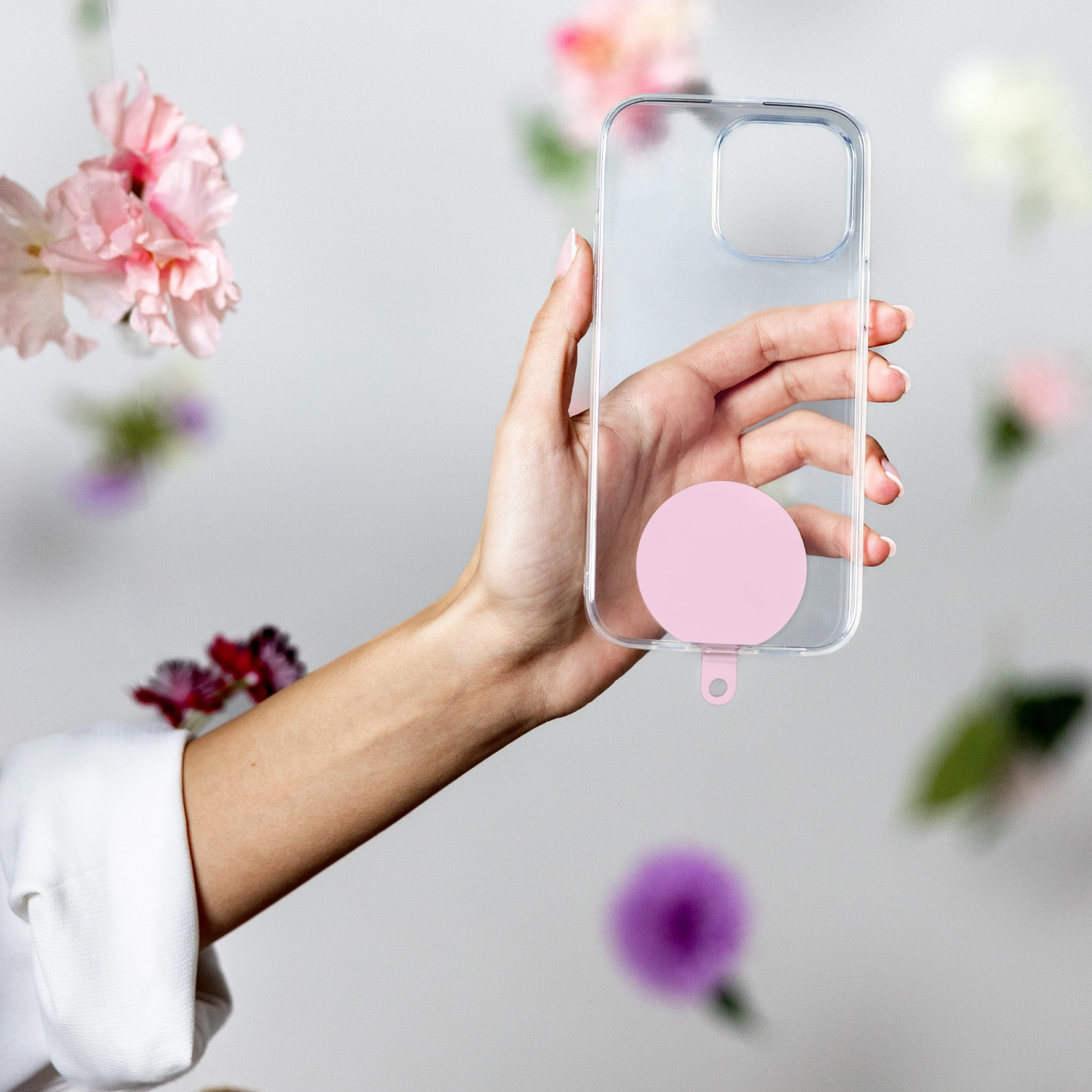 Una mano tiene una custodia trasparente per telefono con un cerchio e un anello rosa. Fiori rosa e viola fluttuano sullo sfondo.