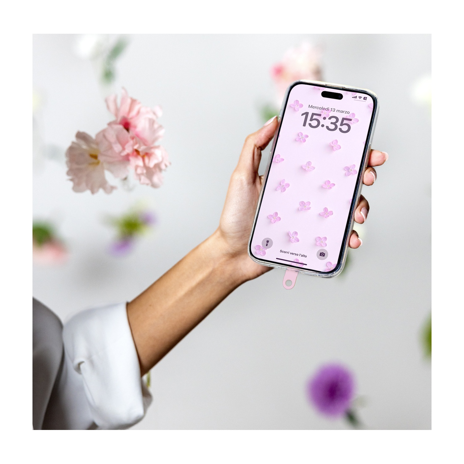 Una mano tiene un telefono che mostra farfalle e ora. Fiori rosa fluttuano sullo sfondo. Il telefono è collegato a un cavo di ricarica rosa.