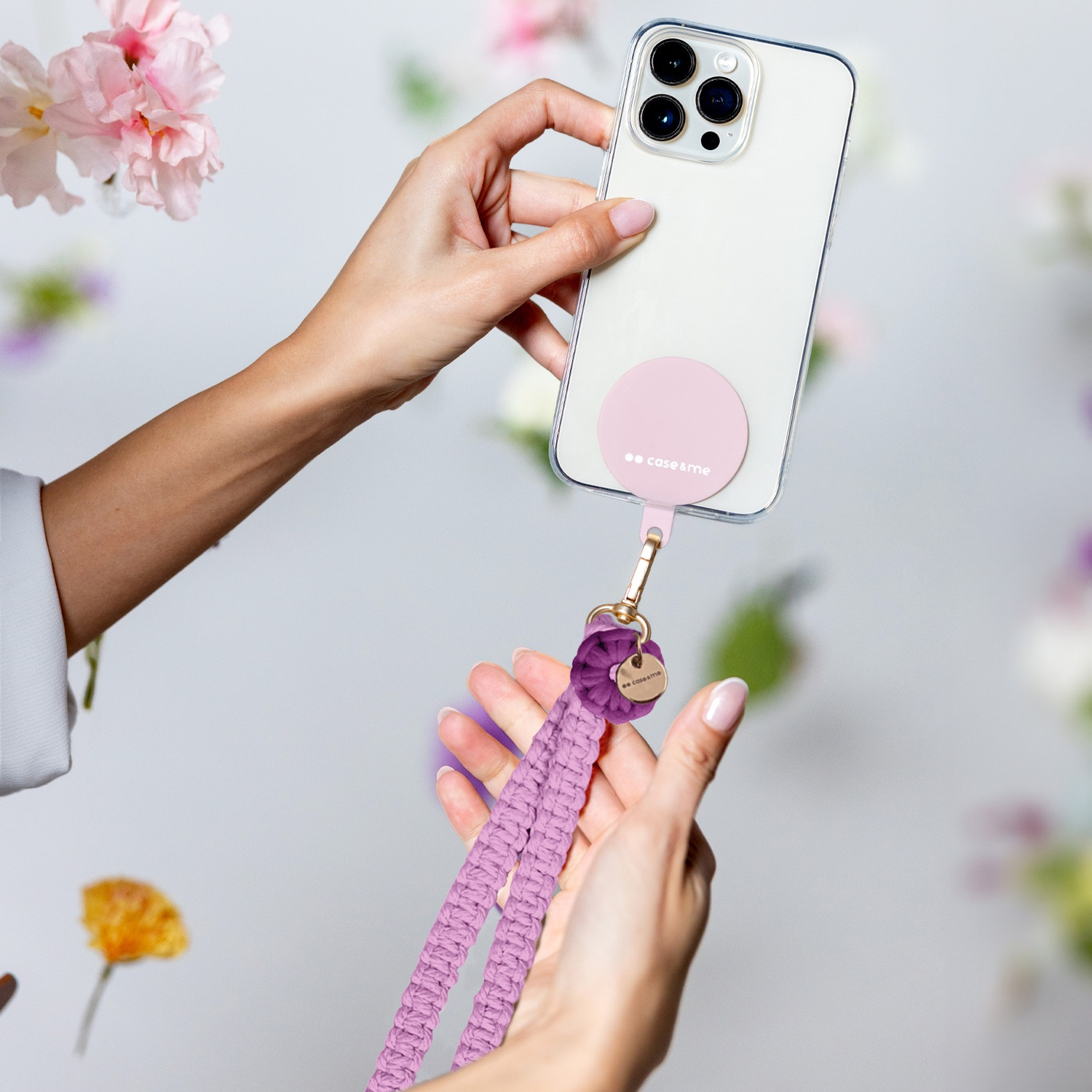 Mani che tengono un telefono con custodia rosa e cordino viola. Fiori sullo sfondo.