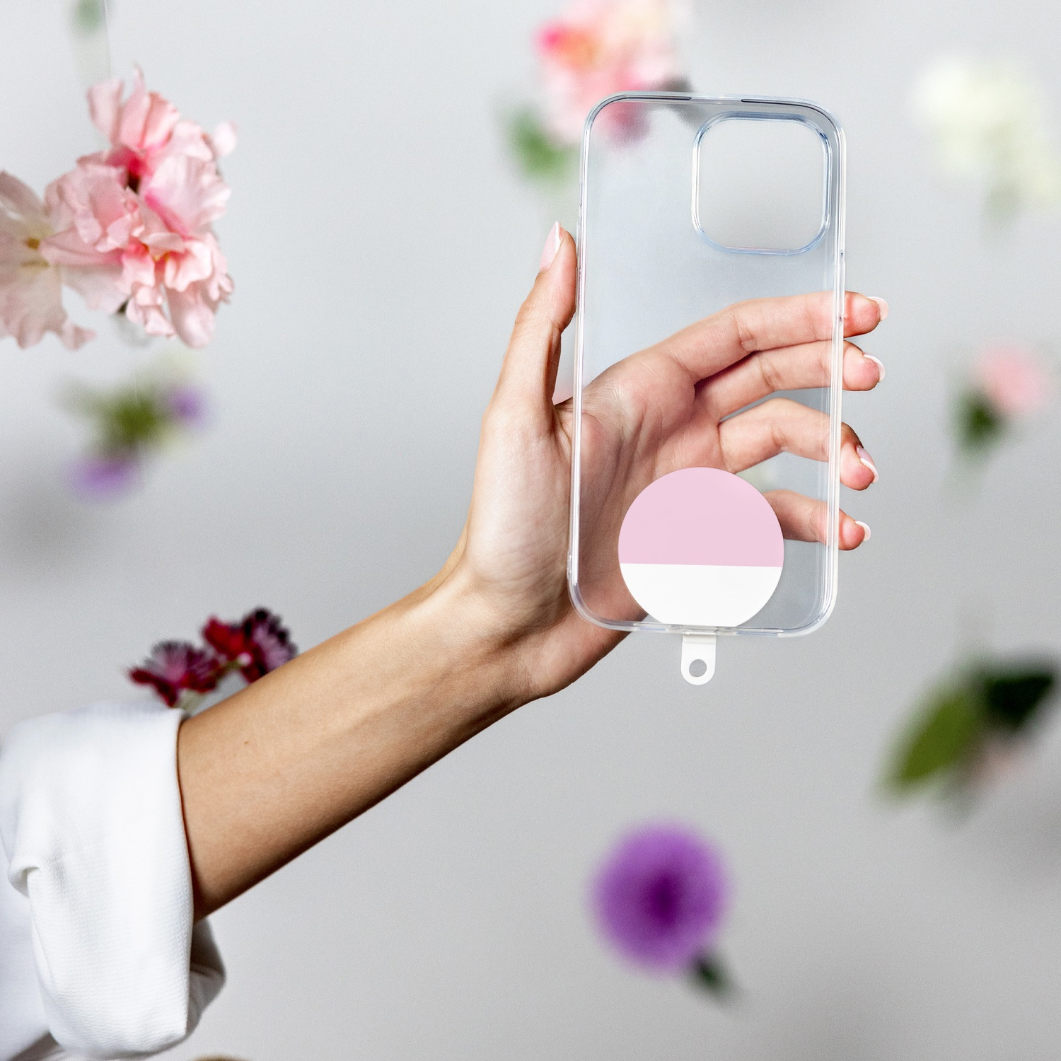 Una mano tiene una custodia trasparente per telefono con un adesivo rosa e bianco. Fiori sullo sfondo sfocato. Manica bianca visibile.