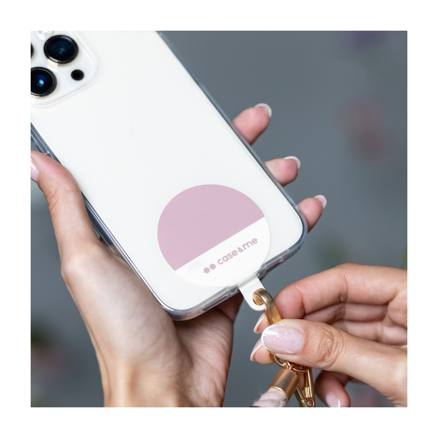 Mani che tengono un telefono con un adesivo a cerchio rosa e un cinturino. Il telefono ha una custodia trasparente. Unghie curate.
