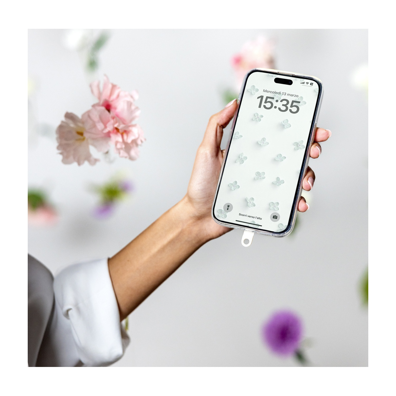 Una mano tiene un telefono con sfondo floreale, circondato da fiori sfocati. L'ora è 15:35. Manica bianca visibile.