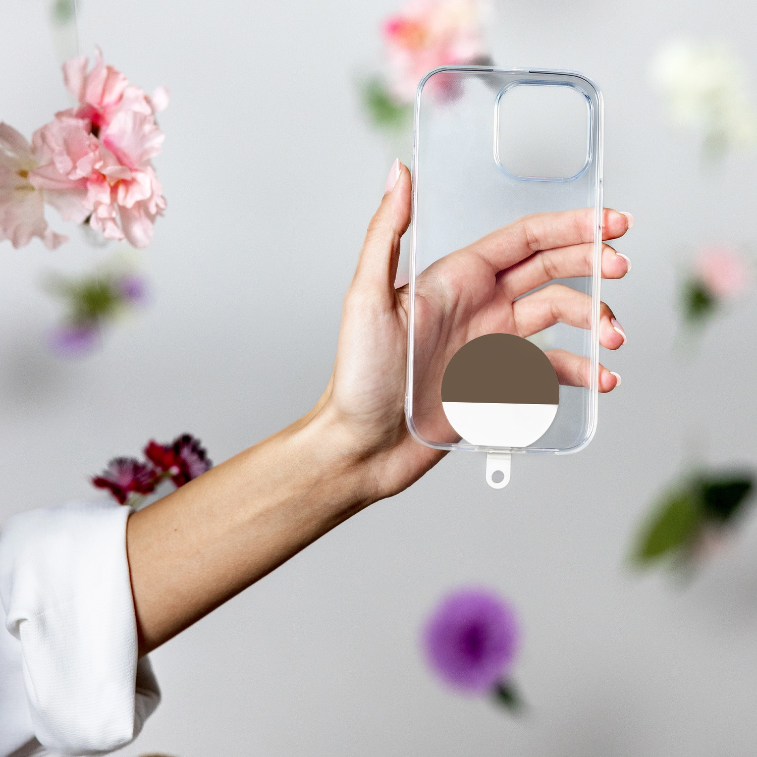 Mano che tiene custodia trasparente per telefono con cerchio marrone/bianco. Fiori rosa e viola sullo sfondo.