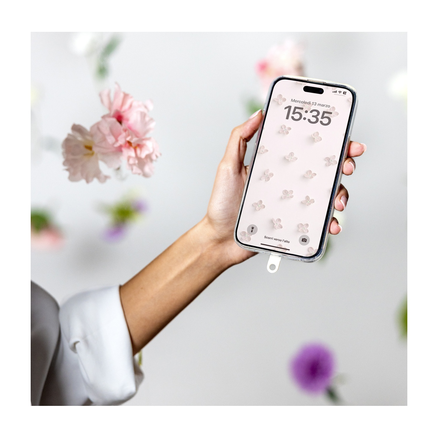 Una mano tiene un telefono con sfondo floreale. Fiori rosa fluttuano sullo sfondo. L'ora sul telefono è 15:35.