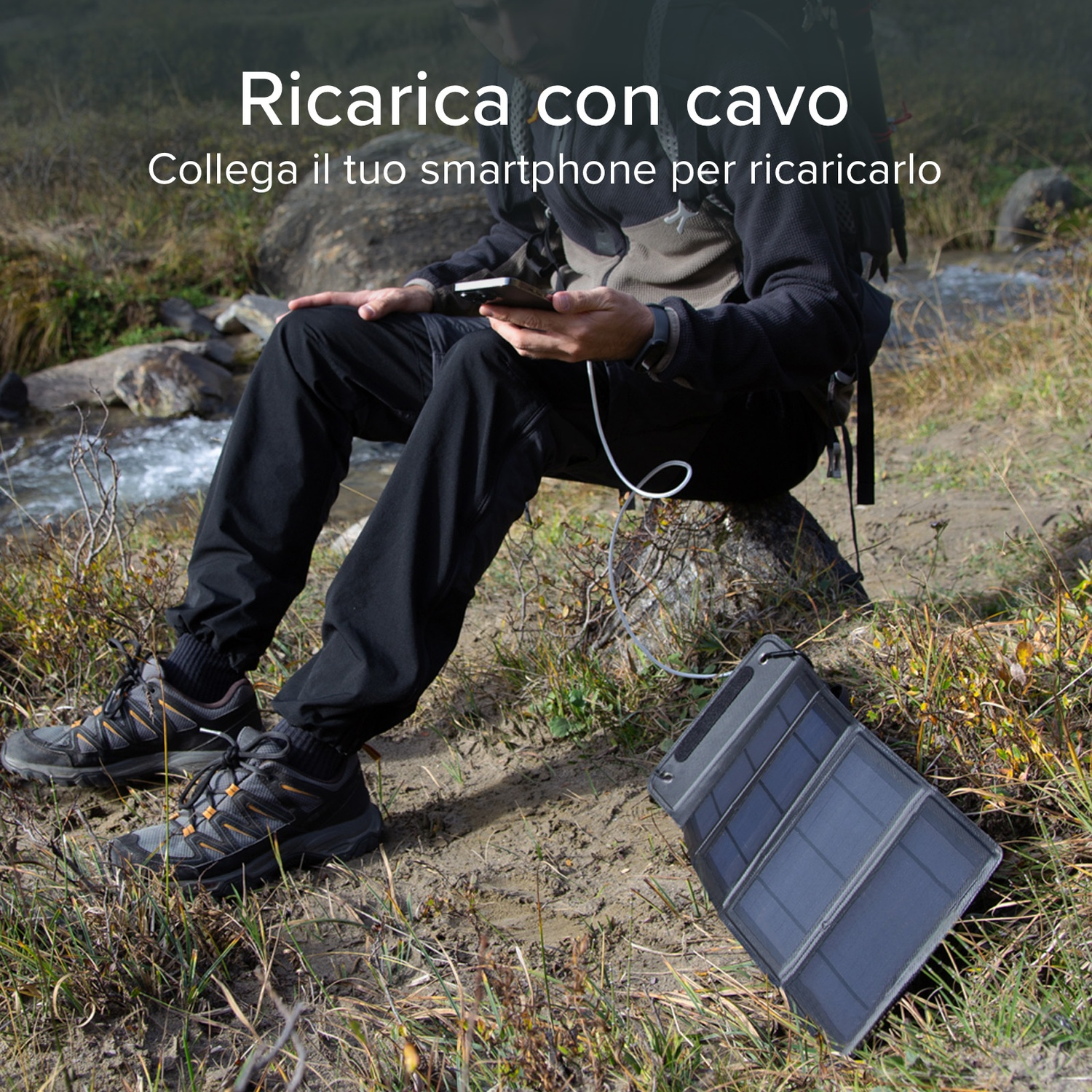 Persona carica telefono con cavo, seduta su roccia vicino a ruscello, pannello solare in primo piano.