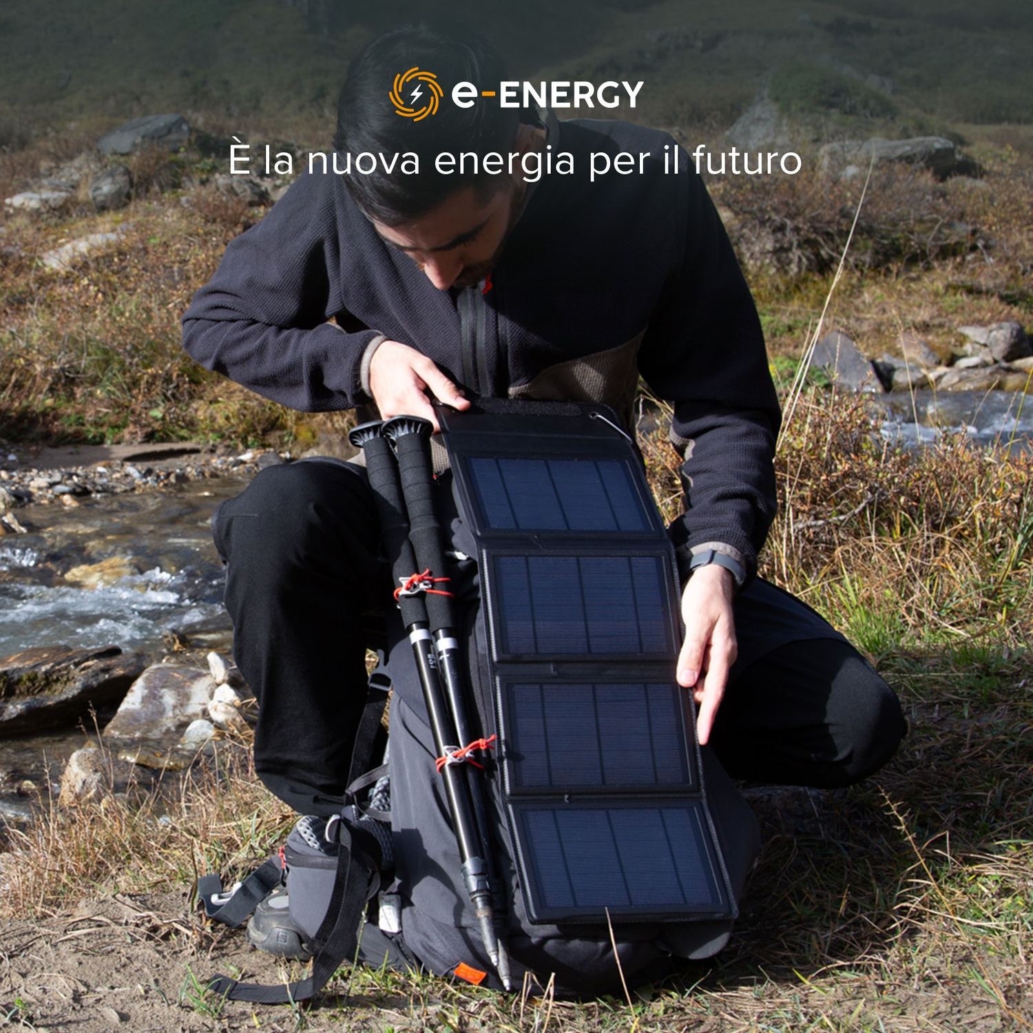 Uomo si inginocchia, apre pannello solare, accanto a zaino e bastoncini. Il ruscello scorre sullo sfondo.