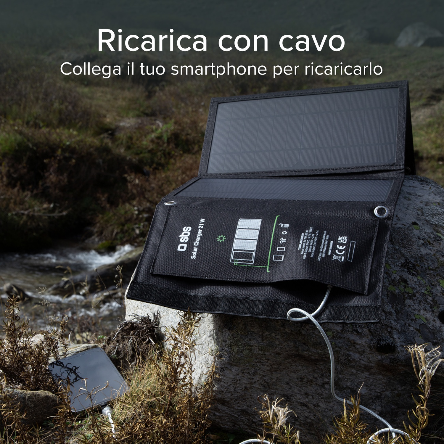 Un caricatore solare nero con un telefono in carica su una roccia, vicino a un ruscello. Il caricatore è aperto, collegato tramite un cavo bianco.