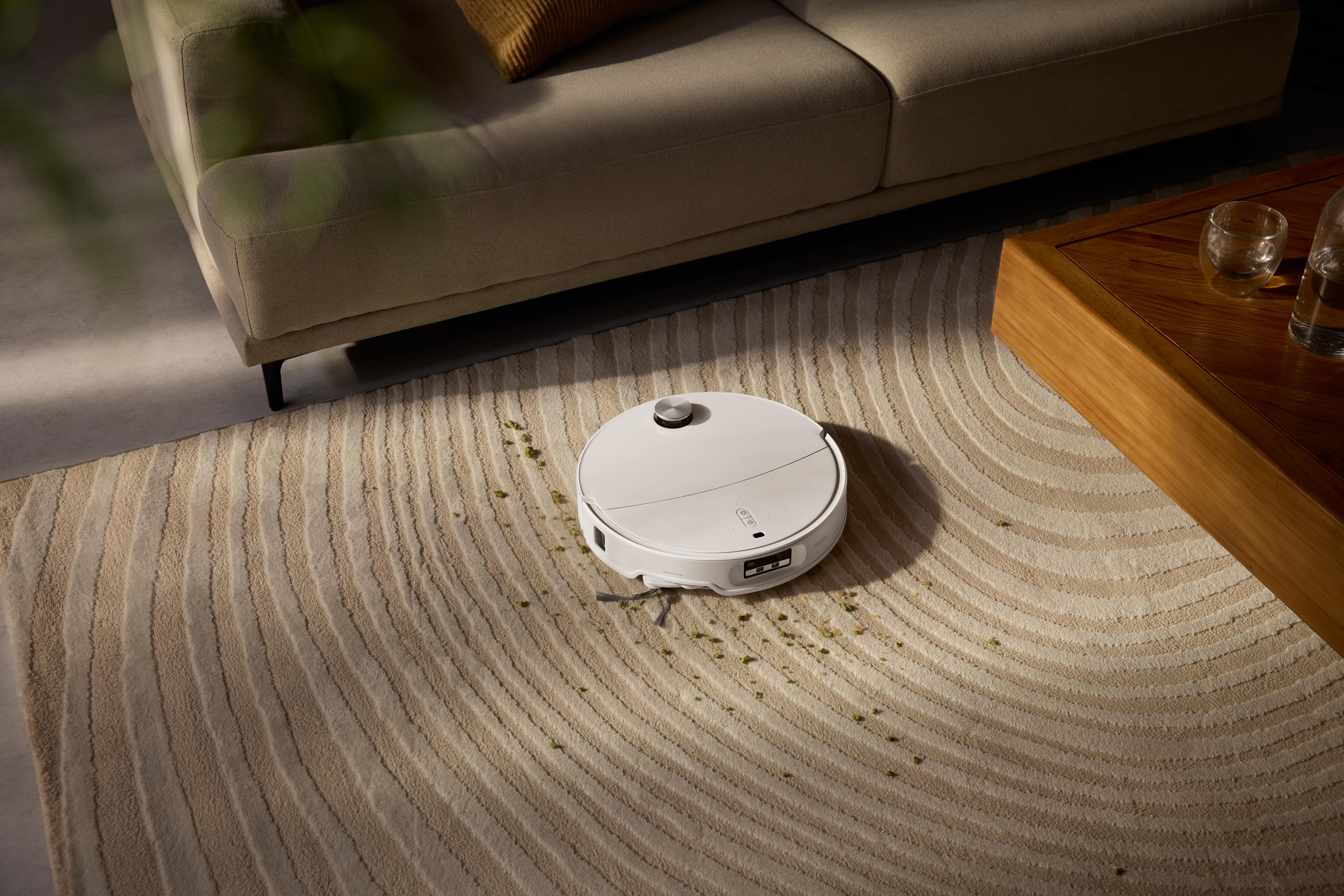 Ein weißer Roboter-Staubsauger auf einem beigefarbenen, gestreiften Teppich, in der Nähe eines Sofas und eines Holztisches. Grüner Schmutz verstreut.