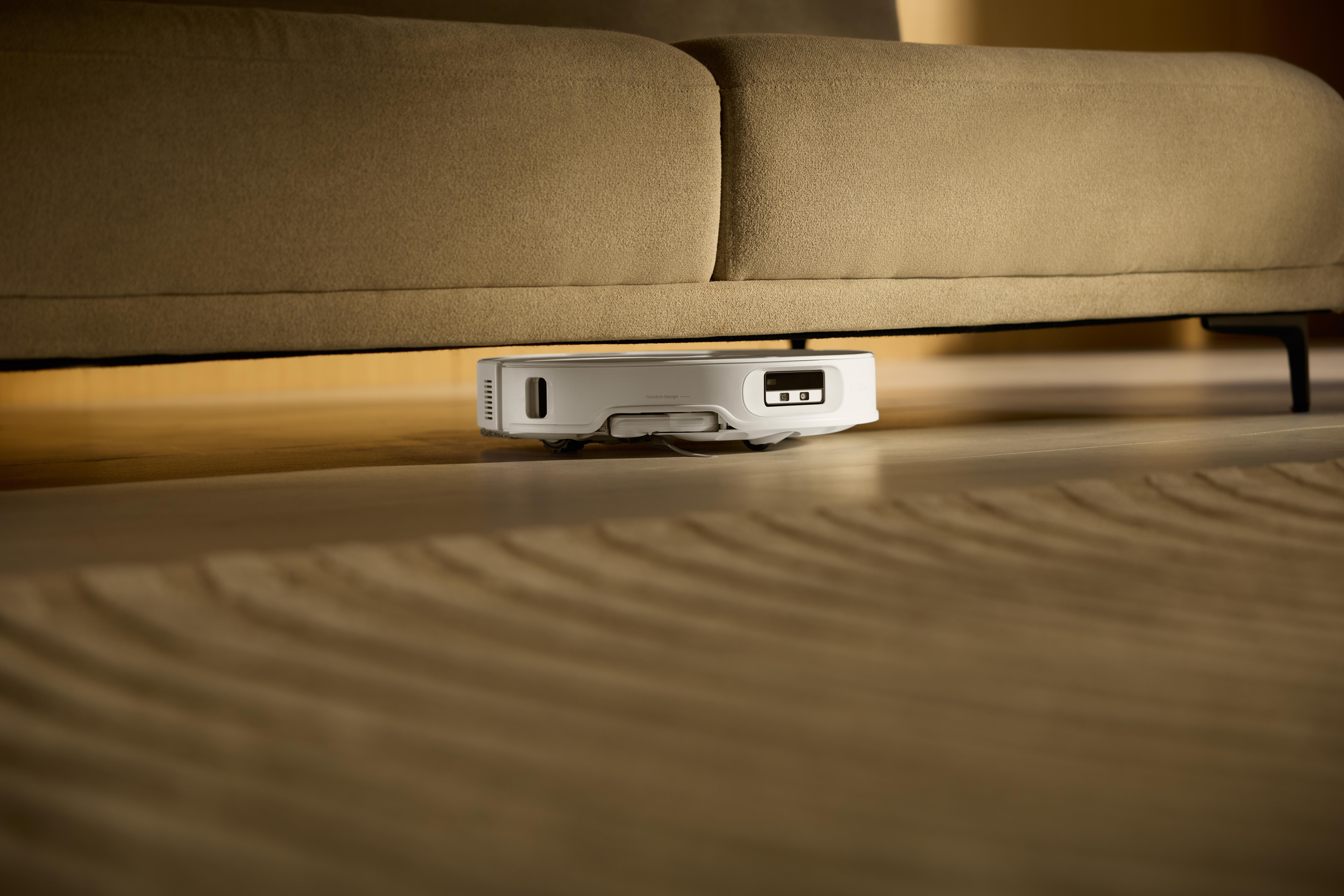 Ein weißer Roboter-Staubsauger unter einem beigefarbenen Sofa. Der Boden ist Holz mit einem hellbraunen Teppich.