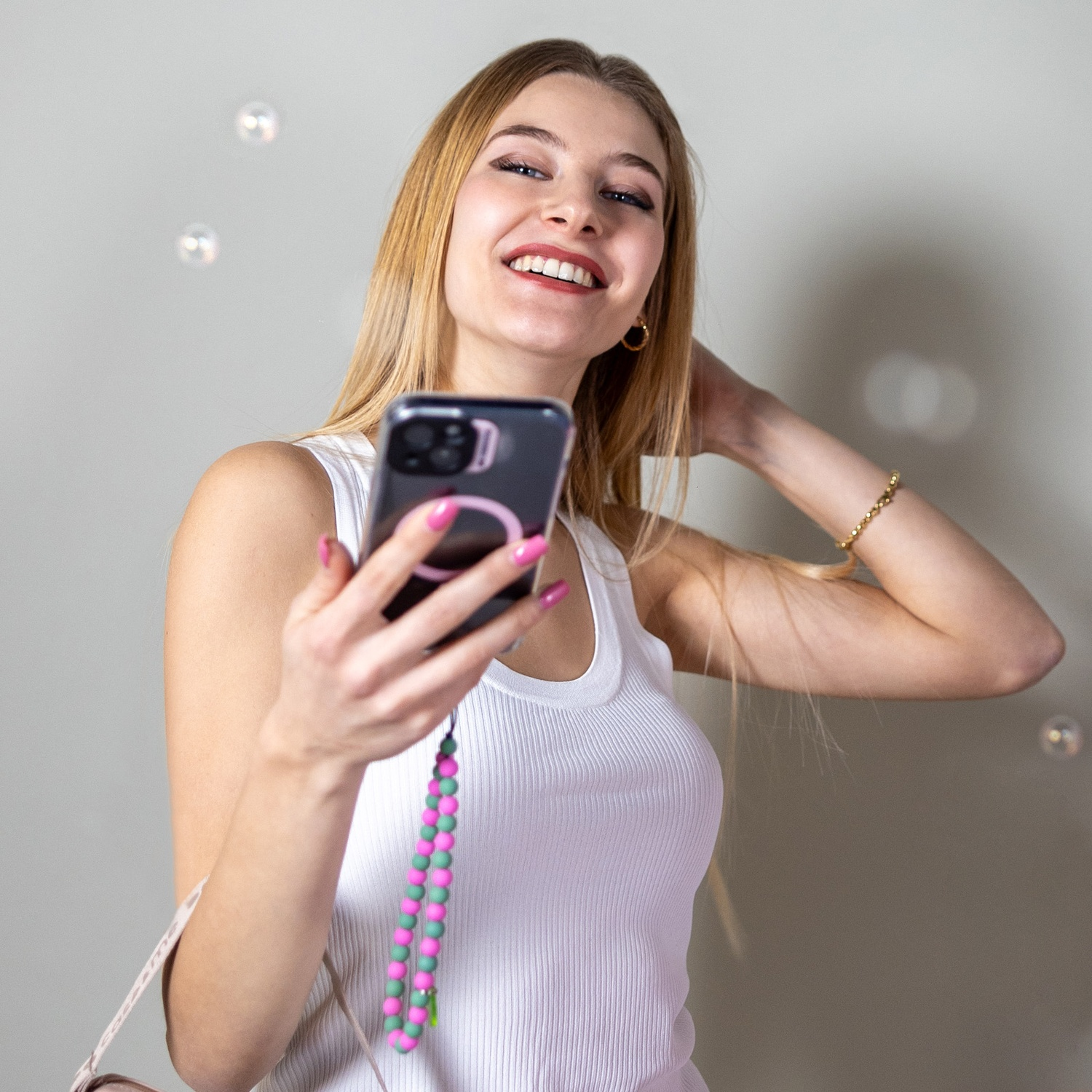 Donna bionda sorride, tiene il telefono, unghie rosa, cinturino di perline verdi/rosa. Canotta bianca, gioielli d'oro e bolle sullo sfondo.