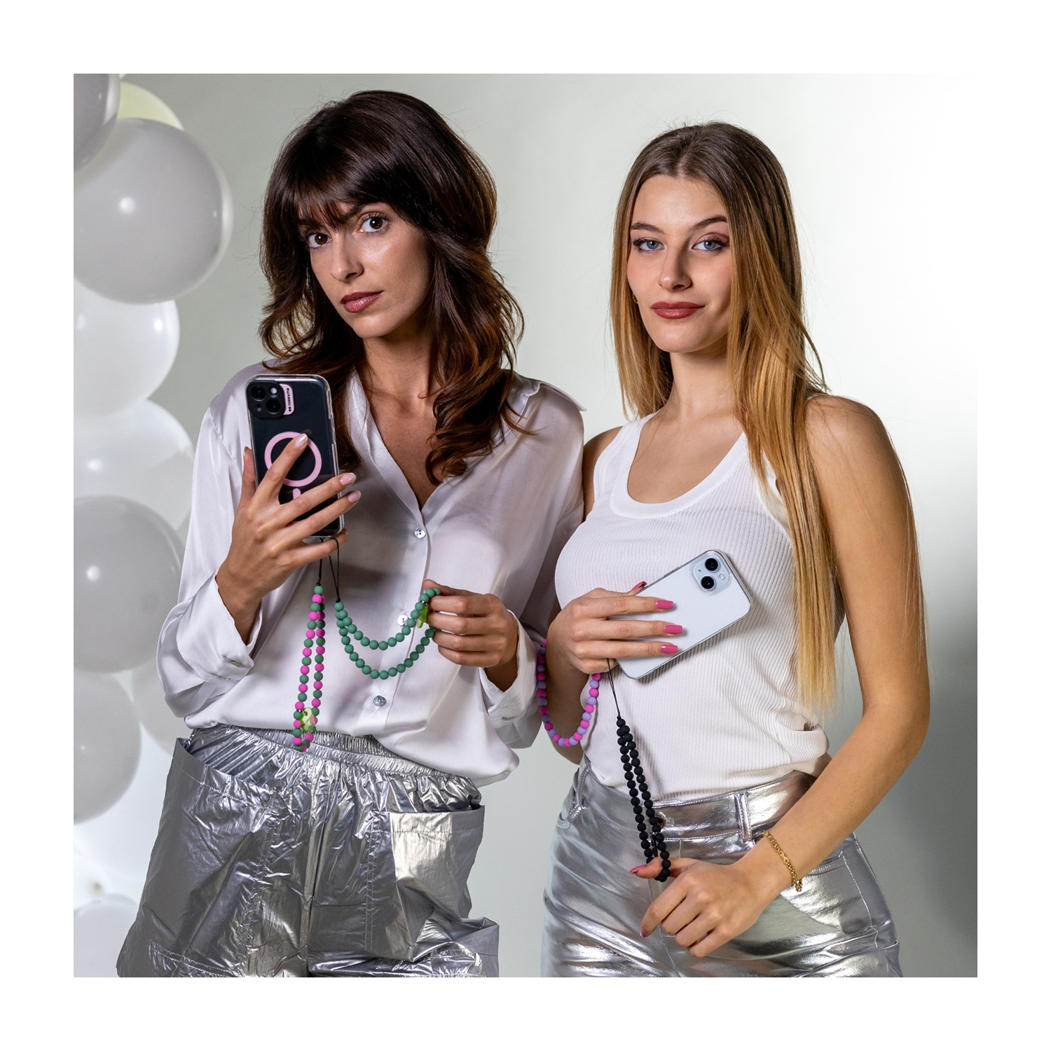 Due donne tengono telefoni con cordini. Una indossa una camicia bianca, pantaloni argentati. L'altra indossa canotta bianca, pantaloni argentati. Palloncini sullo sfondo.