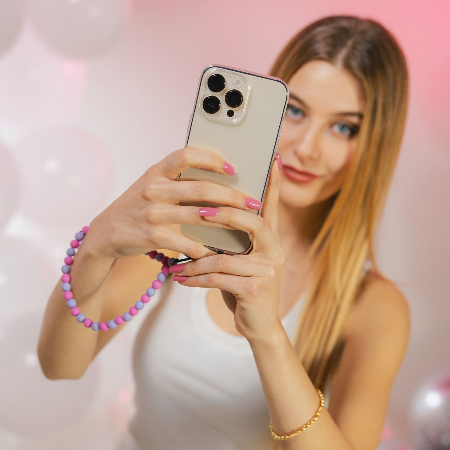 Donna bionda che scatta un selfie con il telefono, indossa un top bianco, un cinturino di perline e unghie rosa, sfondo di palloncini.